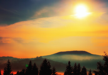 A colorful photo of a mountain landscape at sunset. forest, mountains and bright sun in red clouds