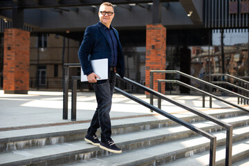 Handsome mature businessman wearing stylish eyeglasses holding laptop computer, standing on the street. Successful business concept