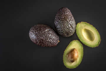 Halved avocado with a stone and two whole ripe avocados on a black background