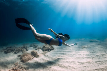 Free diver woman with fins glides underwater over sand in blue ocean.