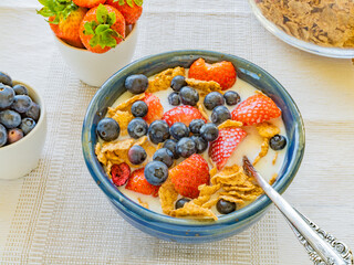 blue bowl with flakes, fresh strawberries and blueberries, milk and small bowls of ingredients on the side