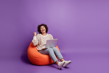 Smiling woman typing on laptop computer rejoice victory raise fist shout sit beanbag on purple wall