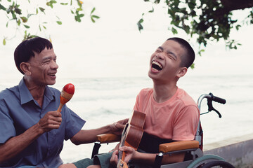 Asian special child on wheelchair is singing, playing music therapy on the beach with parent,Natural sea beach background,Life in the education age of disabled children,Happy disability kid concept.