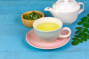 Obraz na płótnie Canvas Moringa Tea in white cup and fresh green leaf on blue wooden background. Moringa oleifera tropical herb healthy lifestyle concept.