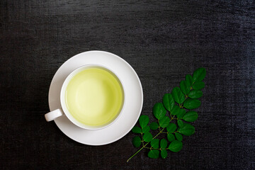 Obraz na płótnie Canvas Moringa Tea in white cup and fresh green leaf on black wooden background, top view. Moringa oleifera tropical herb healthy lifestyle concept.