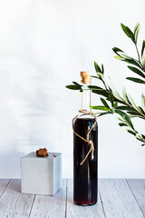 A bottle of strong alcoholic drink with a green olive branch on a light background. Minimalistic still life bottle of homemade liquor or tincture
