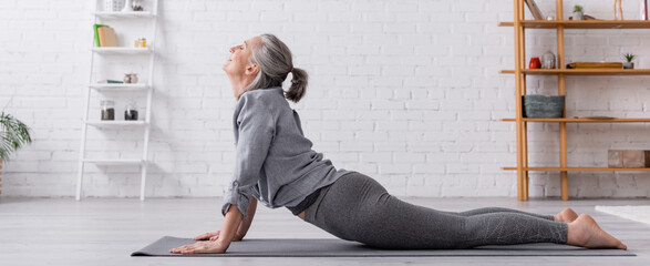 side view pleased middle aged woman in cobra pose practicing yoga on mat at home, banner