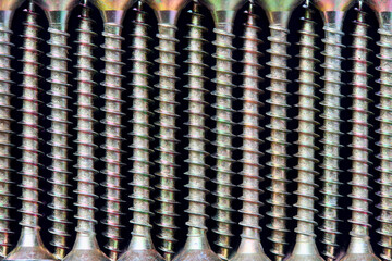 Selective focus golden brass steel wood tapping screws galvanized with rainbow tint lined ornamental macro background pattern, lying in a row over black surface, closeup top view.