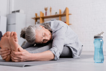 Obraz premium middle aged woman with grey hair stretching on yoga mat near sports bottle