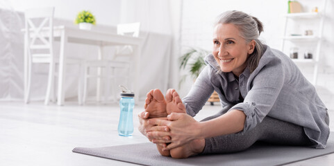 happy mature woman with grey hair stretching on yoga mat near sports bottle, banner