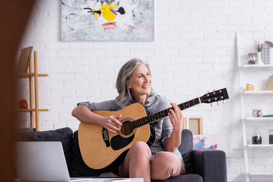 pleased mature woman with grey hair learning to play acoustic guitar near laptop