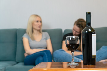 Two young women, a blonde and a brown-haired woman, are sitting on a sofa and talking, in focus in the foreground is a bottle of wine and a poured glass of wine