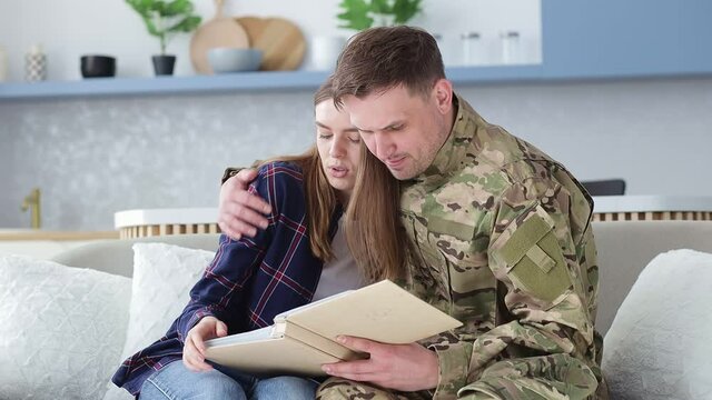 a man hugs a woman while sitting on the couch shows an album with photos from the army with friends with whom he served.
