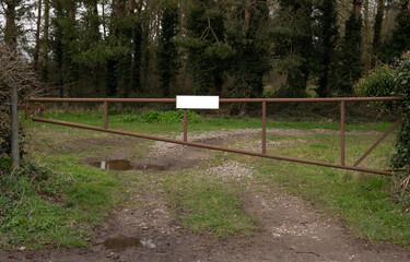 A closed rusty metal bar gate with a blank white board screwed to it ,infront of a farmers field