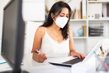 Portrait of busy female entrepreneur in medical face mask sitting at office desk with papers and laptop. Concept of precautions and social distancing in coronavirus pandemic