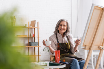 happy middle aged artist holding paintbrush, cup of coffee and using smartphone near canvas and blurred foreground