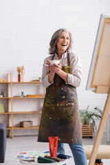 excited middle aged artist holding cup of coffee and paintbrush near canvas