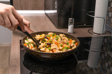 Women's hands are stirring with a spatula pieces of white meat, a variety of vegetables that are...