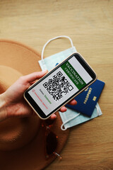 New normal concept. Close up view of female's hold a smartphone with an immune digital health passport. In the background, a sunglasses, protective mask, hat and passport. in a blur.
