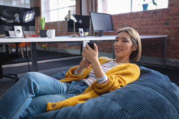 Happy mixed race businesswoman chilling in office relaxing space lying in beanbag using smartphone