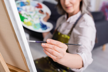 partial view of mature woman holding paintbrush while painting on canvas with blurred foreground