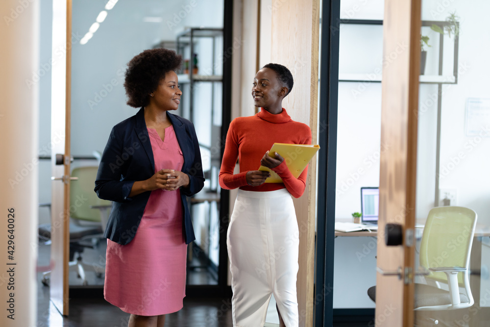 Wall mural Happy african american female business colleagues walking through corridor holding documents talking