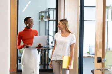 Happy diverse female business colleagues walking through corridor holding documents and talking