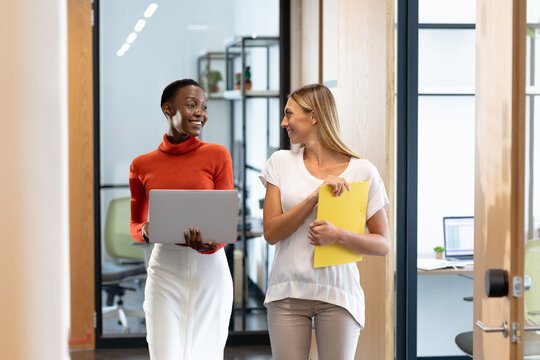Happy diverse female business colleagues walking through corridor holding documents and talking