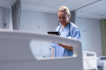 Naklejka na ściany i meble Smiling caucasian female doctor in hospital wearing scrubs and stethoscope using tablet