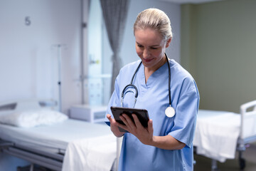 Smiling caucasian female doctor in hospital wearing scrubs and stethoscope using tablet