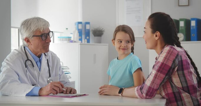 Aged male pediatrician doctor talking to little girl and mother in office of medical clinic.