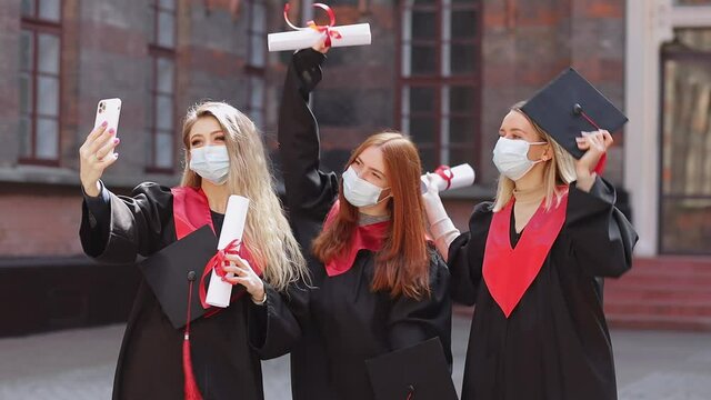 funny girls jumping, dancing, waving diplomas, students in medical masks and black robes make selfie on the phone against the background of the university during quarantine