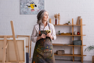middle aged artist in apron holding paintbrush near easel