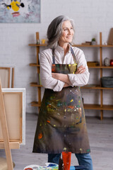 cheerful middle aged artist in apron standing with crossed arms near easel