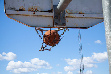 A basketball ball flies into a hoop with a metal net outdoors against a blue sky with white clouds...