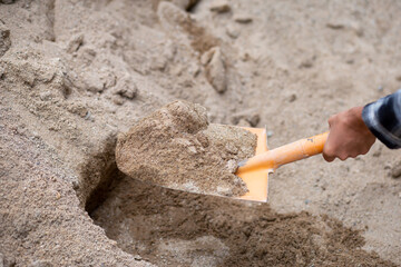 A close-up of the sand in the shovel, the hand of a construction worker shoveling the sand with a shovel. From sandpiles to mix cement on the construction site - obrazy, fototapety, plakaty