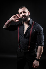 Uomo moro tatuato con la barba in camicia nera e bretelle rosse, porta la mano all'orecchio,...