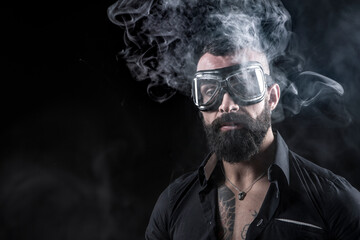 Uomo moro con la barba occhiali da motociclista,  fuma una sigaretta e produce una nuvola di fumo, isolato su sfondo nero