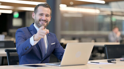 Middle Aged Businessman with Laptop Pointing at Camera 