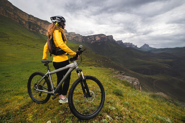 woman on a mountain bike stands high in the mountains looks at the mountains and the sky in the...