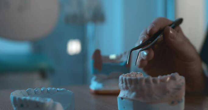 Close-up a gypsum model of the human lower jaw while a hand of the medical worker is holding dental tweezers with ceramic olay putting it on the gypsum tooth model of a human jaw.
