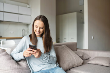 Serene young woman holds smartphone sitting on the couch at home, chatting online with friends, spend leisure time in social media, texting, scrolling news feed, shopping online