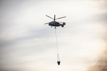 Fire fighting helicopter silhouette with bambi bucket for сarrying water to put out a massive building city fire, process of put out a large blaze bush fire wildifre, aerial firefighting with chopper