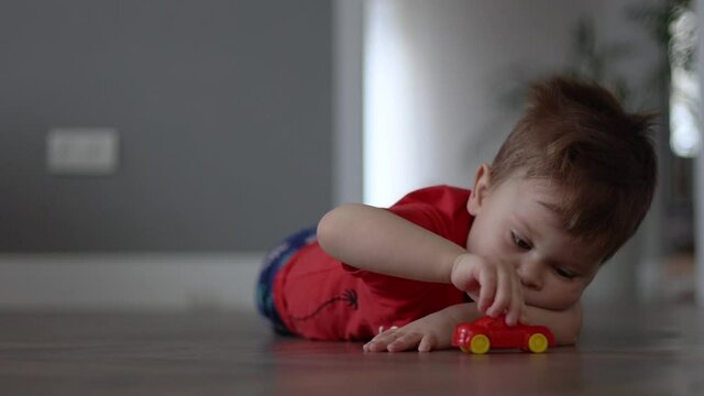 sad little kid in red t-shirt lies on the wooden floor and plays with toy car on the floor at home