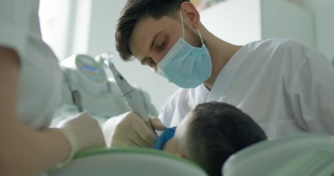 A little boy is laying in the dental chair and having dental treatment by the male dentist who is using the dental tool.