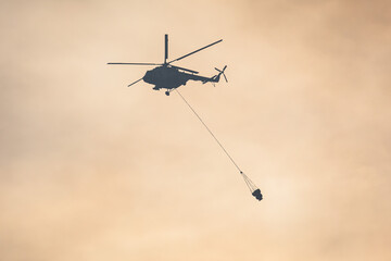 Fire fighting helicopter silhouette with bambi bucket for сarrying water to put out a massive building city fire, process of put out a large blaze bush fire wildifre, aerial firefighting with chopper
