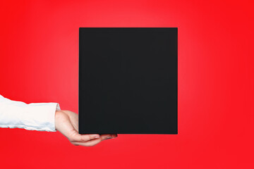 A square black box with an copy empty space for text and design in the hand of a young woman in a white shirt on a red background. Board, blank, template, mockup, layout for a slogan or inscription - Powered by Adobe