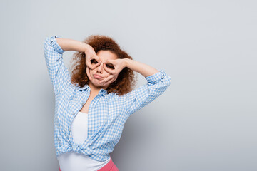 Fototapeta premium curly woman having fun while imitating binoculars with hands on grey background