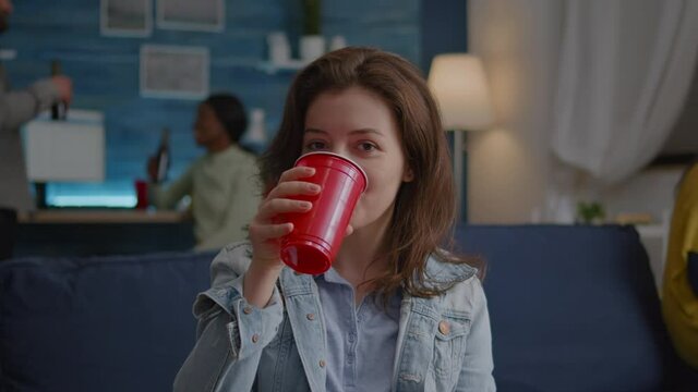 Portrait of woman sitting on sofa similing at camera while drinking beer late at night during holiday party. In background multi-ethnic friends gathering together for celebrating birthday