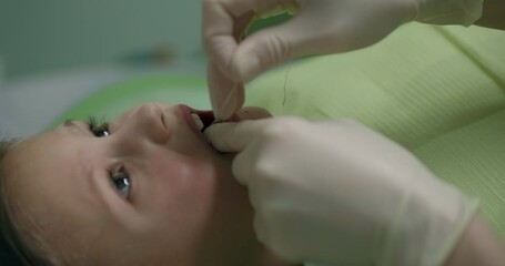 Close-up a little girl is laying in the dental chair with an open mouth while a dentist is using dental floss for cleaning her teeth.